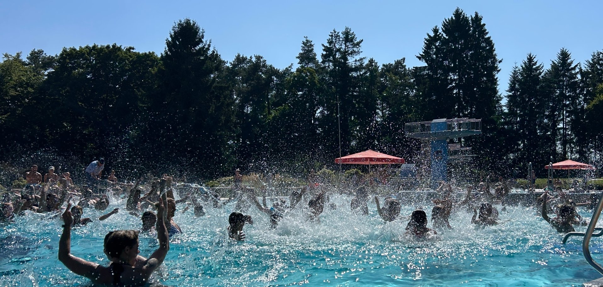 Freibad Miesau Ein turkisblaues Schwimmbad mit Badegästen in im Hintergrund grüne Bäume und blauer Himmel.