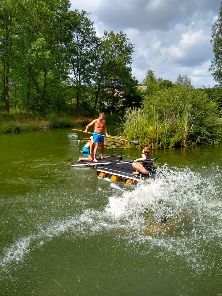 Zwei Menschen in Badebegleidung auf einem Floß und ein weiterer sitzender Mensch auf einem Floß. 