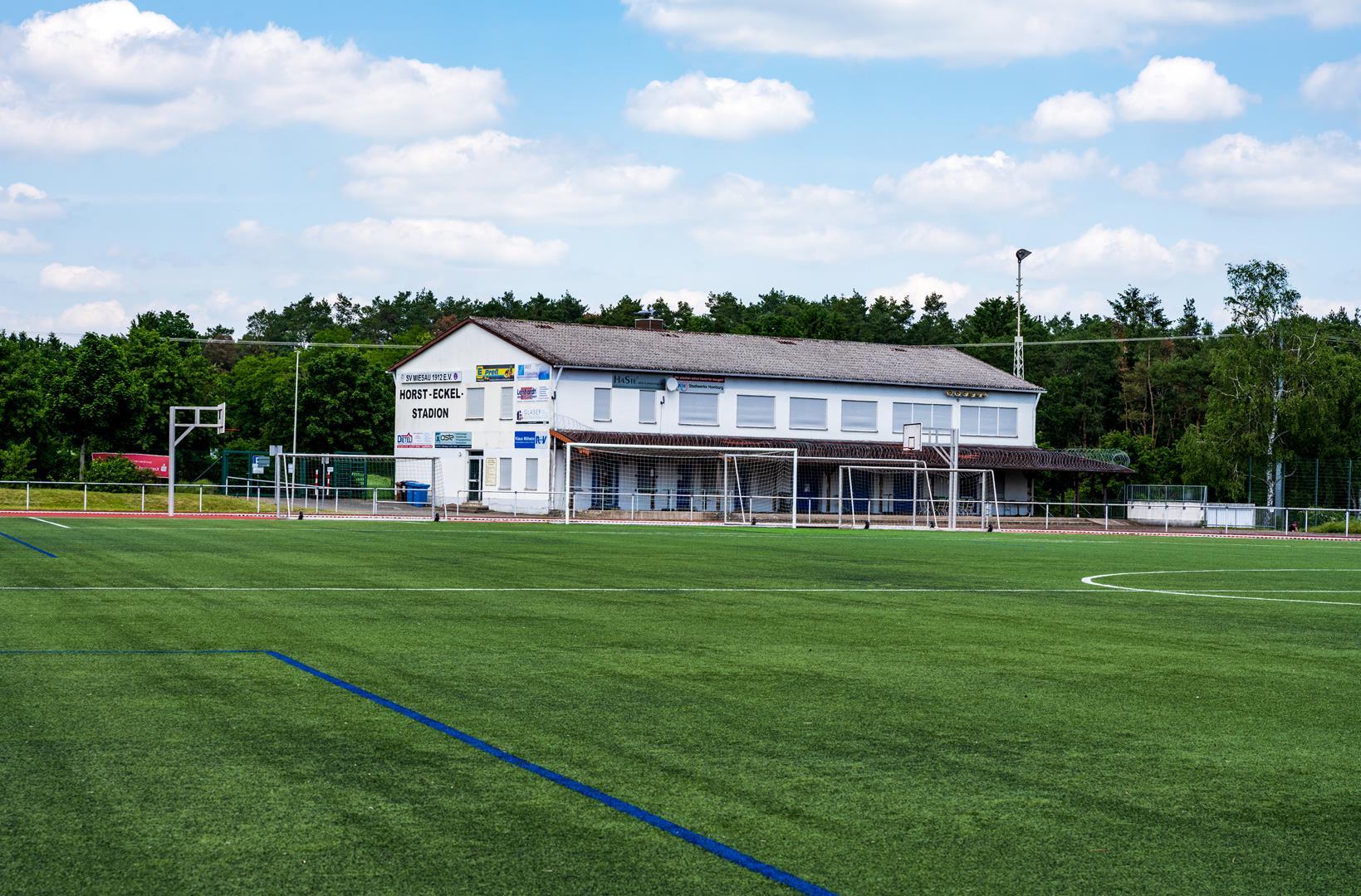 An einem Fußballfeld befindet sich ein weißes Haus, auf dem Horst Eckel steht.