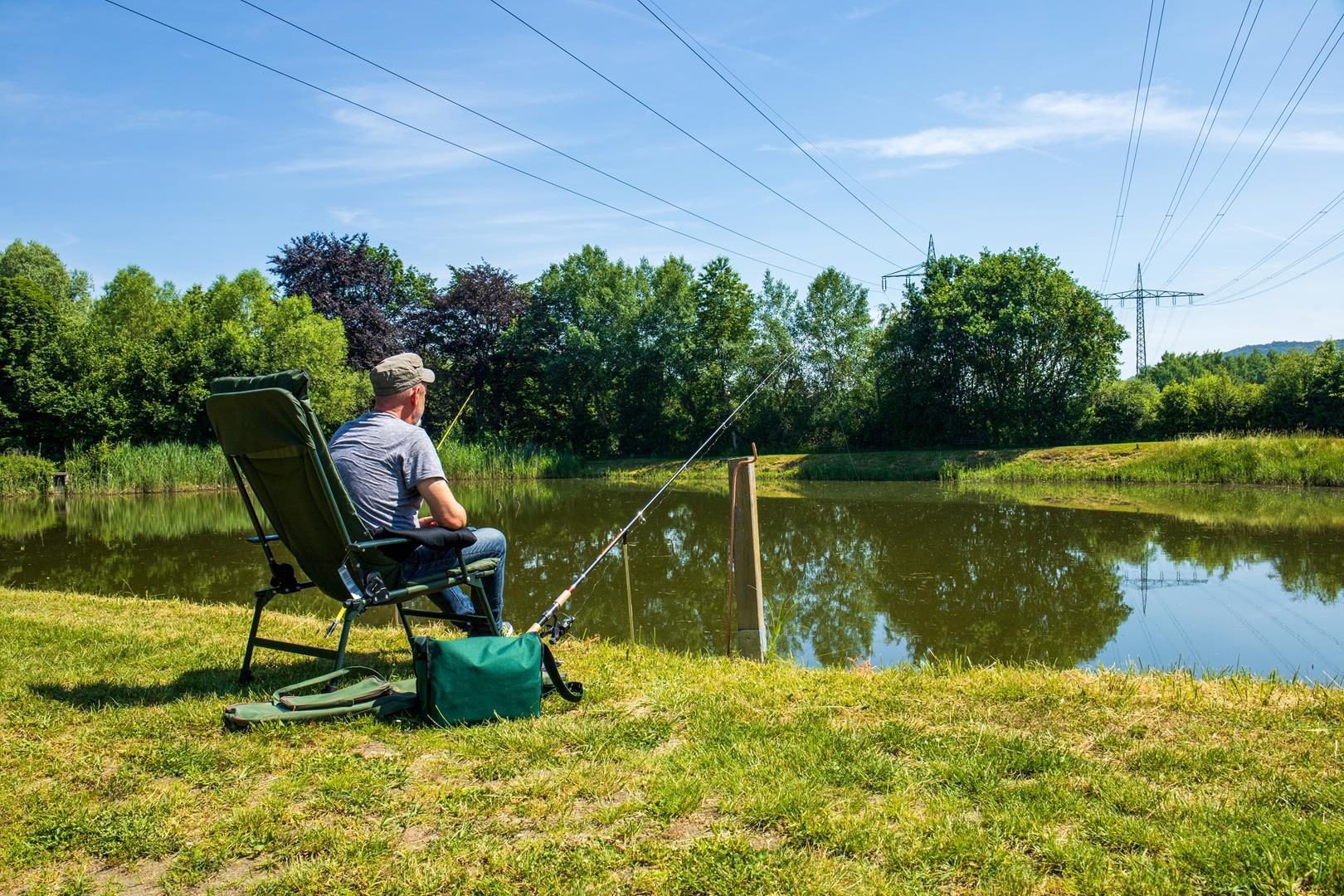 Ein Angler in einem Campingstuhl an einem See sitzt und in einer Hand eine Angel hält. Neben ihm eine weitere Angel  