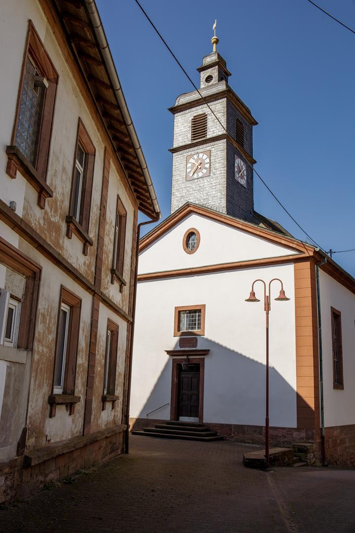 Auf der linken Seite ein renovierungsbedürftiges Haus, das den Blick auf die Frontseite der Lambsborner Kirche lenkt.