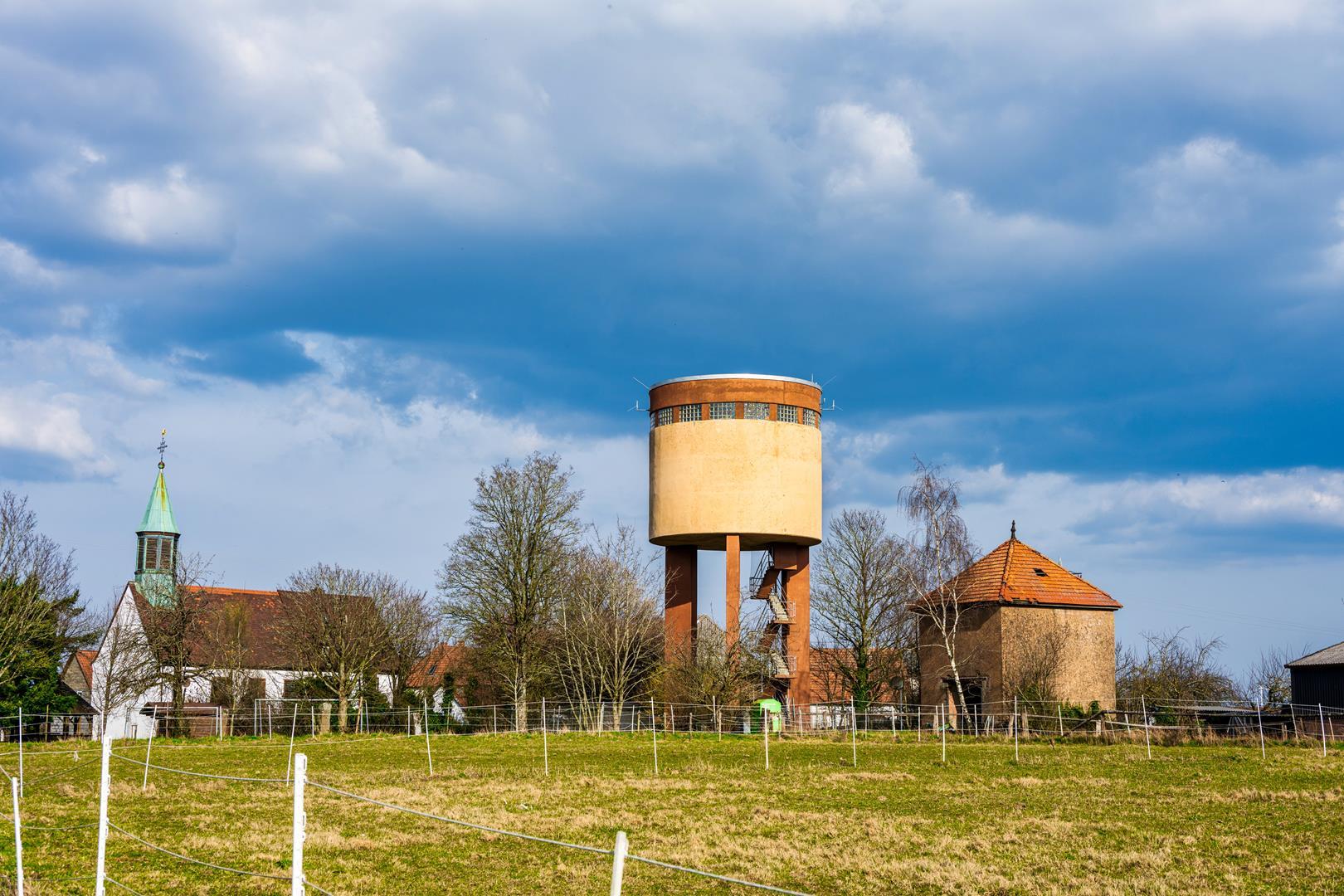 Wasserturm auf einer Wiese. Im Hintergrund ein Dorf.
