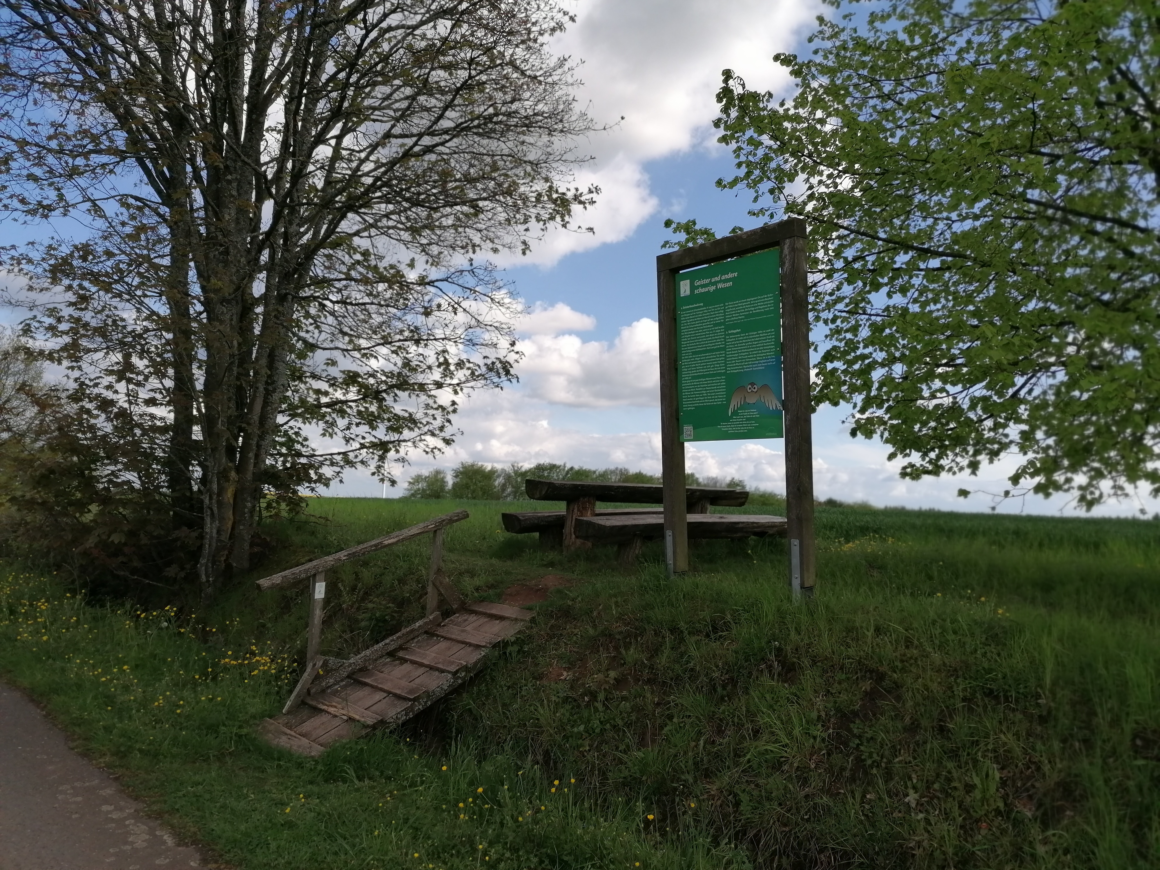 Tisch mit zwei Bänken unter einem Baum zu dem man über eine kleine Holzbrücke erreicht