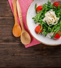 Kulinarische Angebote Salat mit Rucola und Tomaten in einer weißen Schüssel. Neben der Schüssel liegen zwei Holzlöffel auf einer rot weiß karrierten Serviette.