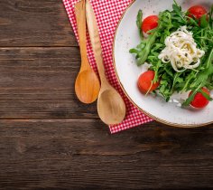 Kulinarische Angebote Salat mit Rucola und Tomaten in einer weißen Schüssel. Neben der Schüssel liegen zwei Holzlöffel auf einer rot weiß karrierten Serviette.