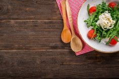 Kulinarische Angebote Salat mit Rucola und Tomaten in einer weißen Schüssel. Neben der Schüssel liegen zwei Holzlöffel auf einer rot weiß karrierten Serviette.