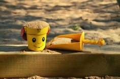 Spielplatz Ein gelber Eimer mit Gesicht und eine gelbe Schaufel mit Sand auf einem Holzbalken.