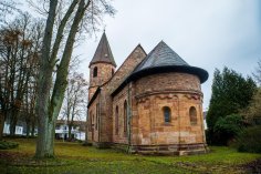Zu sehen ist eine Kirche aus Sandstein mit einem abgerundetem Chor, dem Kirchenschiff und dem Kirchturm.  Die Kirche steht auf einem gepflasterten Platz mit Bäumen. Im Hintergrund sind Häuser zu sehen