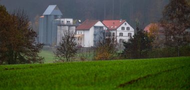 Teilweise verputzte Sandsteingebäude, an das sich ein weiteres Gebäude  anschließt. Dahinter befinden sich Silos