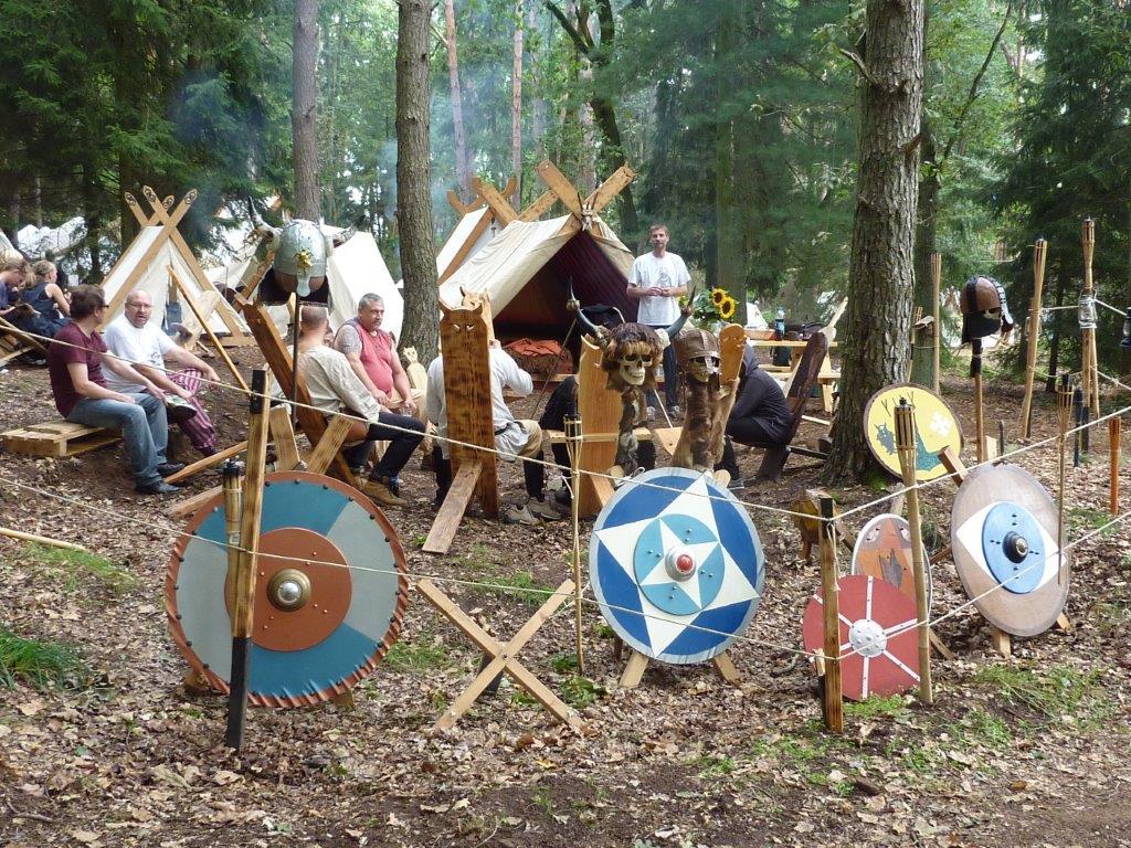 Schilde im Vorderrund. Dahinter eine Gruppe mit Menschen im Kreis. Im Hintergrund weiße Zelte.