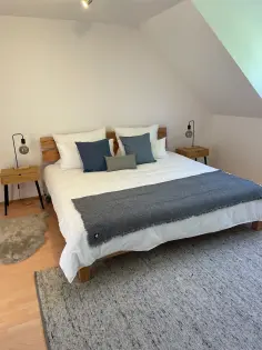 Doppelbett aus Holz mit weißen Bettzeug  und blauen Deko-Kissen sowie einer blaue-grauen Tagesdecke. Daneben stehen zwei Nachttische aus Holz