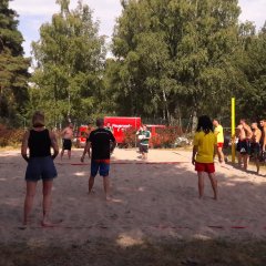 Fünf Menschen stehen im Sand des Vollyballspielfelds vor einem Netz . Hinter dem Netz stehen weitere fünf Menschen.  Andere stehen am Rand des Spielfelds und schauen den spielenden zu.
