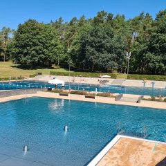 Zu sehen sind zwei Swimmingpools im Freibad Bruchmühlbach-Miesau. Im vorderen Pool, der L förmig  ist, befinden sich drei Masagedüsen. Im Rechten Eck des Bildes ist eine rote Rutsche. Am Rand des hinteren Pools stehen zwei Sprungtürme. Hinter dem Pool stehen eine reihe von Bäumen.